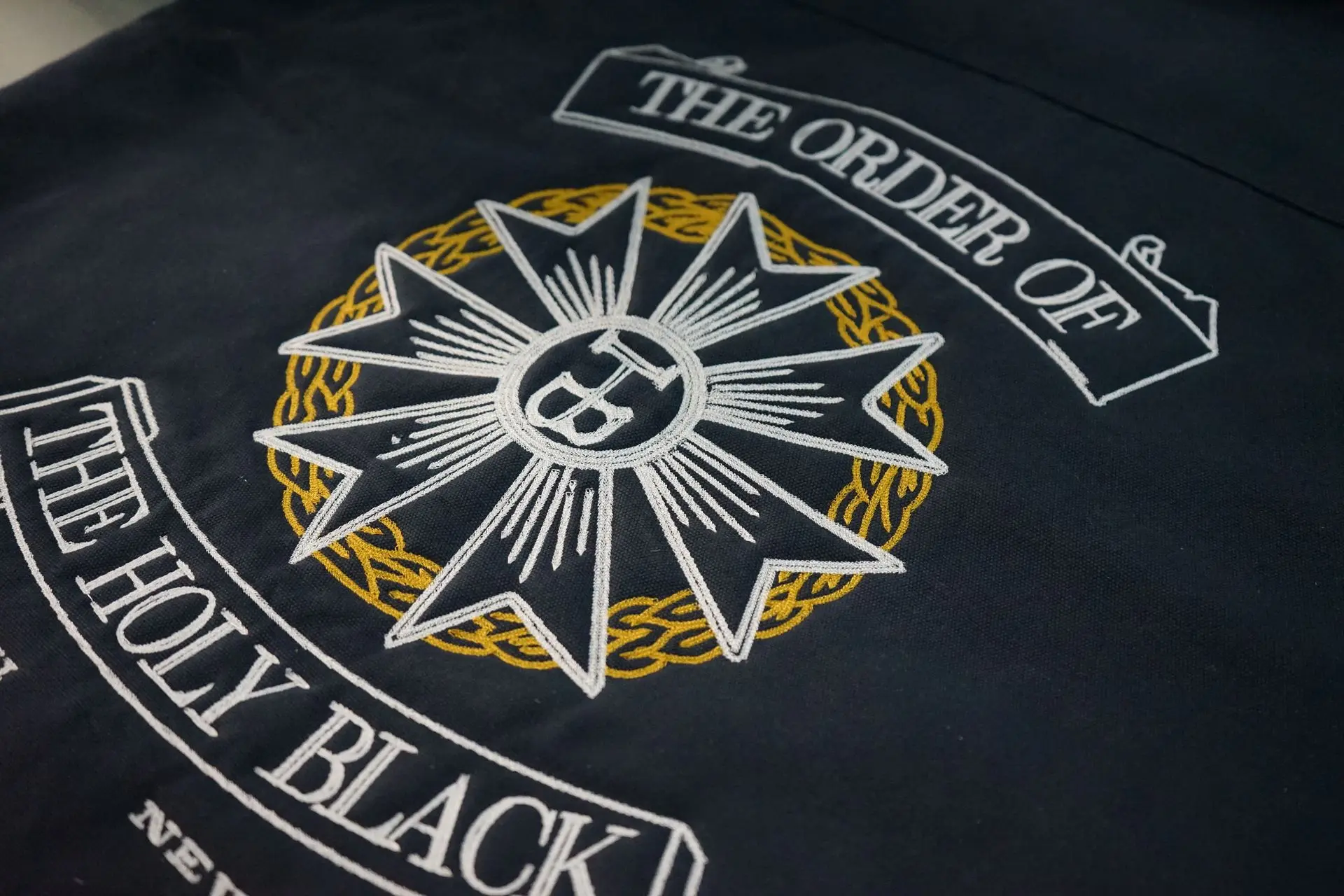 Intricate embroidery of 'The Order of The Holy Black' emblem on fabric showcases exquisite craftsmanship. The design features a radiant star with a barber's straight razor at its heart, encircled by golden laurel wreaths, symbolizing prestige and tradition in grooming excellence.