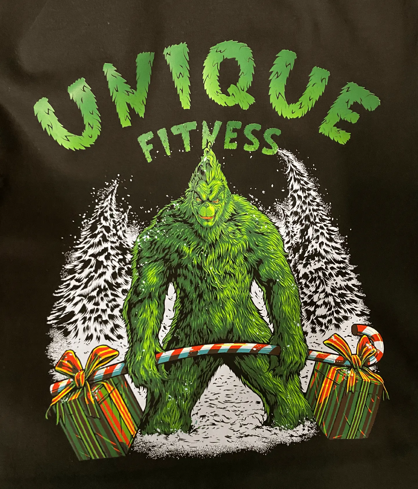 A vibrant and whimsical Unique Fitness holiday-themed shirt design featuring a green Bigfoot character in a snowy landscape, holding a candy cane and surrounded by gift boxes. This eye-catching graphic illustrates the brand's fun and unique approach to fitness and community engagement.