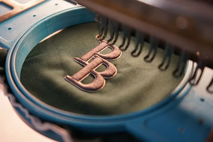 Close-up of an embroidery machine creating a detailed 'BB' monogram on green fabric, showcasing the precision and quality craftsmanship in personalized apparel.