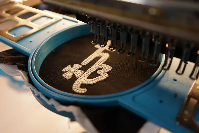 An embroidery machine meticulously crafts a 'HB' monogram on black fabric, showcasing advanced textile customization. This image highlights precision in personalized apparel creation, ideal for branding and bespoke fashion.
