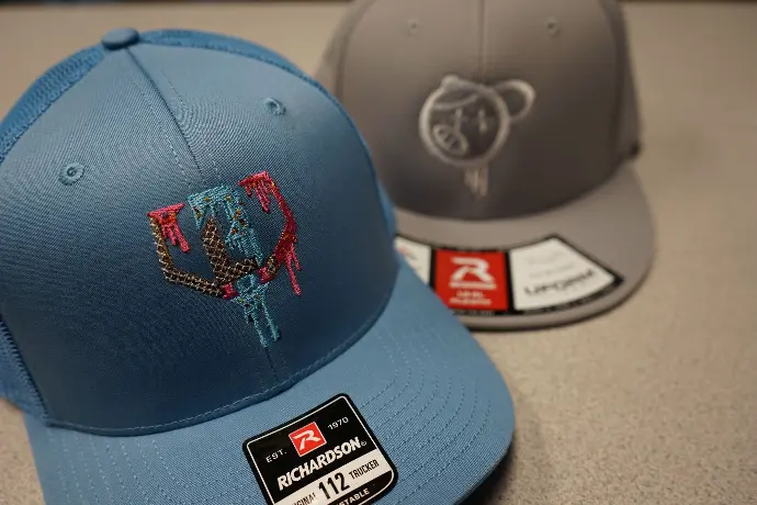 Two Richardson brand caps, one in vibrant blue featuring a detailed, colorful embroidered logo, and the other in grey with a more subdued design. These caps exemplify high-quality craftsmanship and the versatility of custom embroidery in promotional headwear.