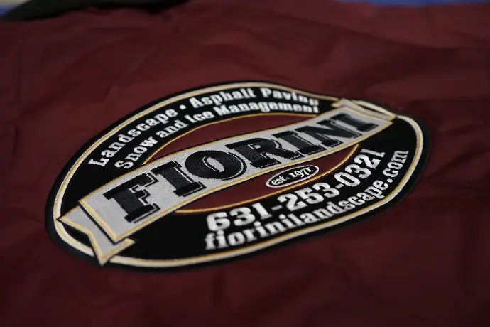Close-up of a high-quality embroidered patch for Fiorini Landscape & Asphalt Paving, showcasing their services in landscape, asphalt paving, snow, and ice management. The detailed embroidery features the company name, 'Florin,' prominently in gold on a maroon and black background, emphasizing professionalism and reliability.