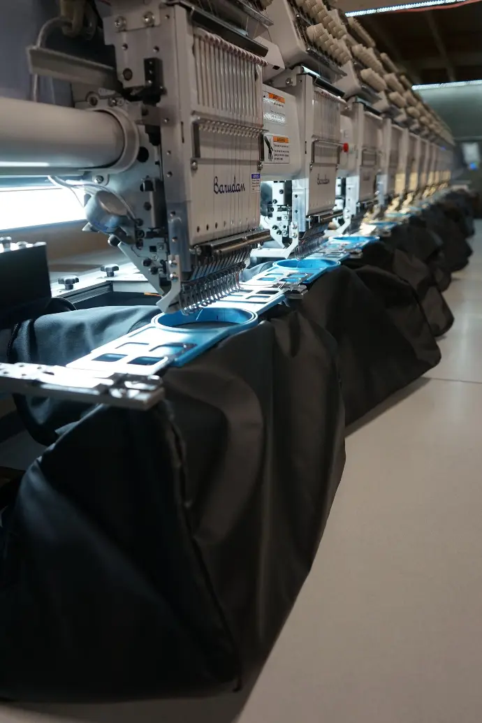 A row of embroidery machines at work on multiple garments, showcasing the scalability of custom embroidery for business and personal apparel. This image represents the efficiency and quality control in producing large orders of branded merchandise.