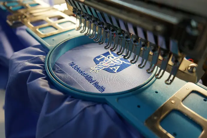 An embroidery machine meticulously crafts the logo of the 'FSA - The School of Arts in Suffolk' on a blue fabric, showcasing the detailed insignia and motto. This image highlights the precision and custom branding capabilities for educational institutions.
