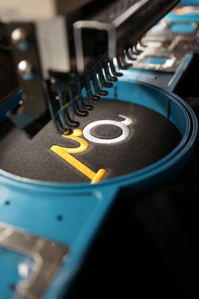 Close-up view of an embroidery machine crafting a vibrant 'IO' monogram on fabric, highlighting the precision and customizability of modern textile technology. This image showcases the intricate details and vibrant color contrasts achievable in personalized apparel.