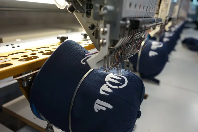 A close-up view of an embroidery machine precisely stitching logo onto a navy blue cap, showcasing the detailed craftsmanship involved in customizing promotional hats. This process highlights the machine's capability to produce crisp, durable text and designs for branded merchandise.