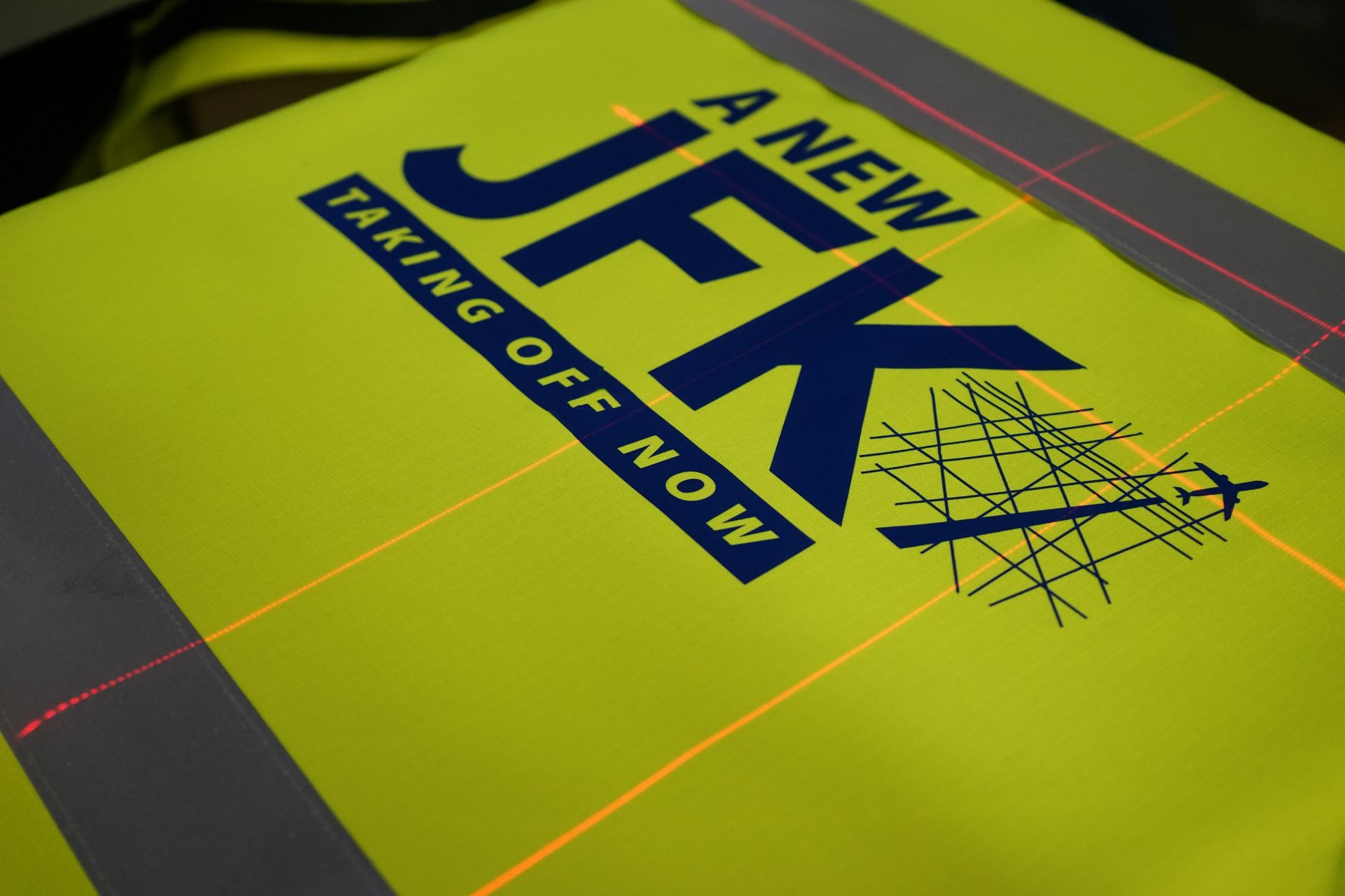 A striking neon green safety vest is adorned with the phrase 'A NEW JFK TAKING OFF NOW' in bold navy blue text. The vest features a lively graphic of flight paths and an airplane silhouette, representing the significant advancements and updates unfolding at JFK Airport.