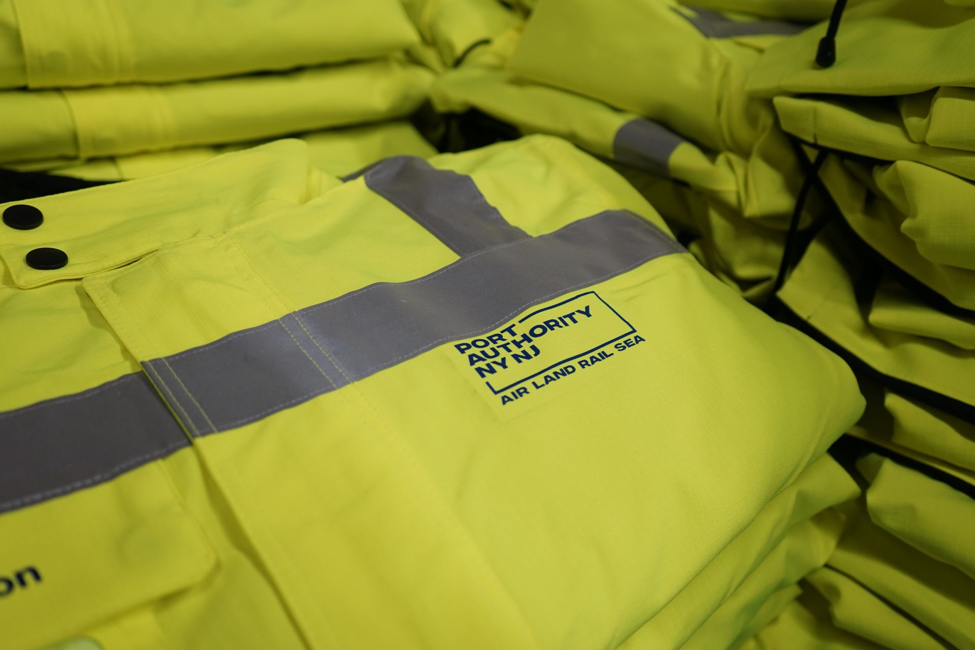  close-up view of high-visibility safety vests featuring the 'Port Authority NYNJ' logo in bold text. The vibrant neon yellow vests, underscored with reflective silver stripes for enhanced visibility, are designed for optimal safety in various transport settings including air, land, and sea operations.