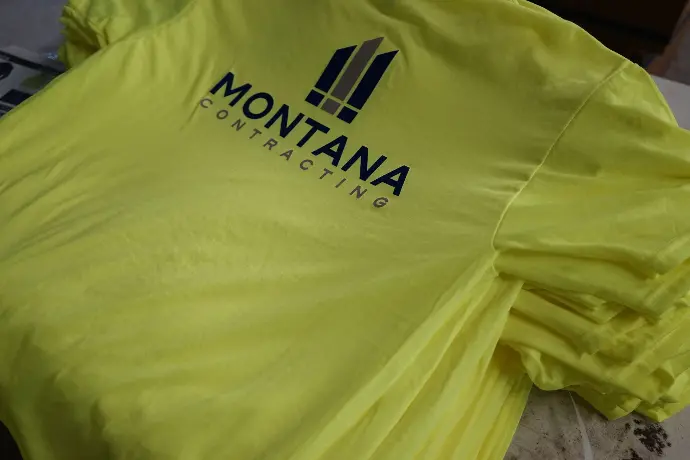 A safety shirt in a striking shade of yellow showcases the 'MONTANA CONTRACTING' name. Its bright color is tailored for high visibility, ensuring safety on the construction site, while the navy blue logo underscores the firm’s reputation for quality and a steadfast commitment to secure working conditions.